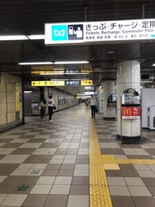 駅構内の通路と、出口・方面を示す頭上の案内板を写した縦位置の写真。